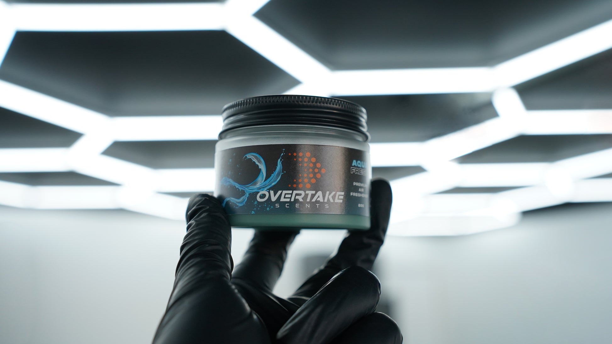 Aqua Fresh gel car air freshener held in black-gloved hand at car detailing studio with hexagonal ceiling lights — Overtake Scents