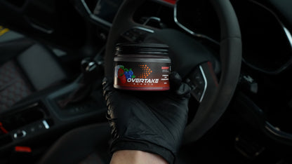 Berry Chill gel car air freshener held in black-gloved hand inside car, steering wheel and red-accented interior visible — Overtake Scents