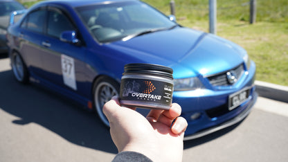 Black Crush gel car air freshener held in hand in front of blue sedan with race number 37 at car event — Overtake Scents