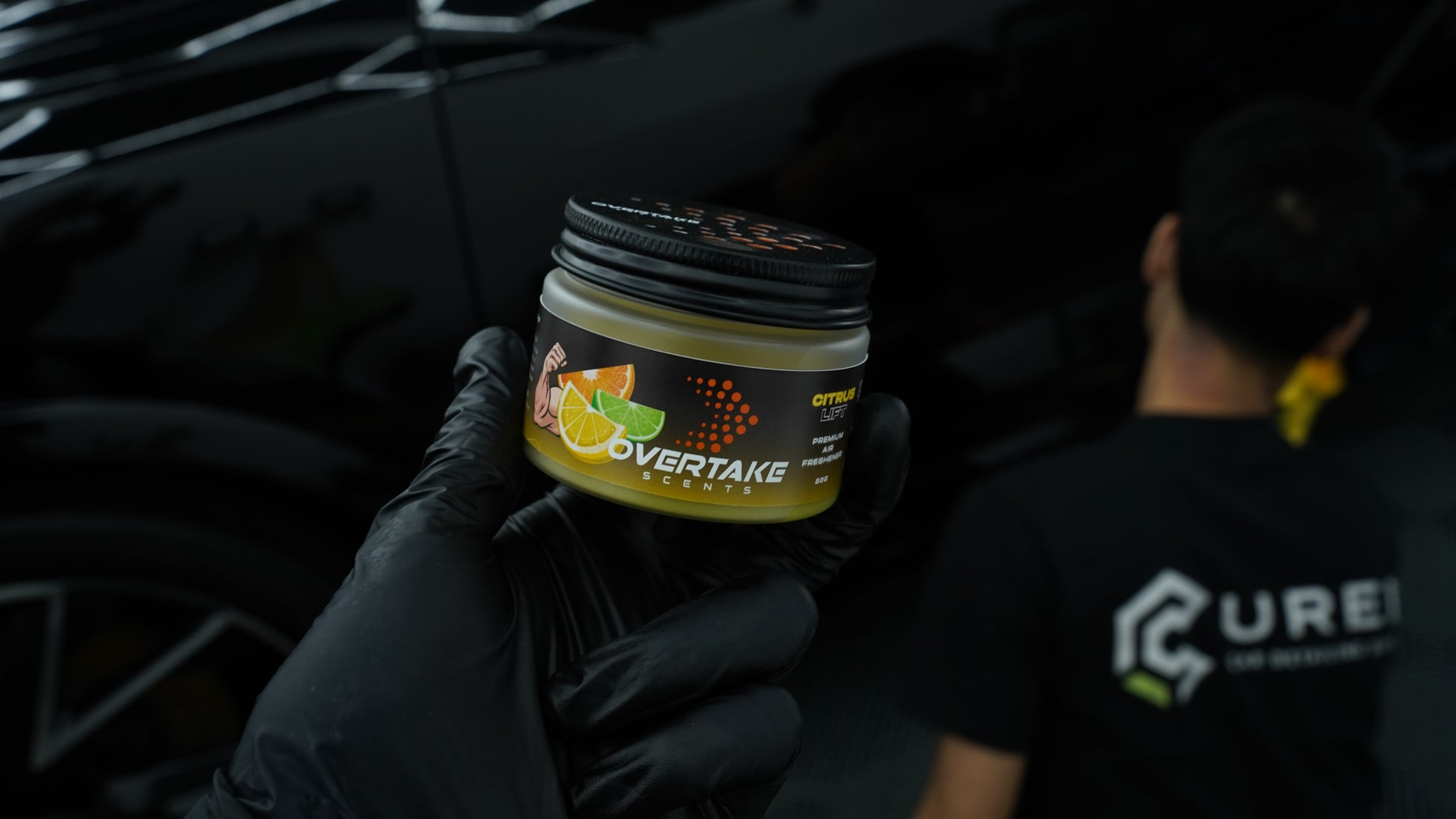 Citrus Lift gel car air freshener held in black-gloved hand at car detailing studio, detailer in background — Overtake Scents