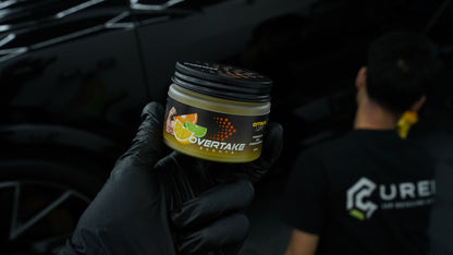 Citrus Lift gel car air freshener held in black-gloved hand at car detailing studio, detailer in background — Overtake Scents