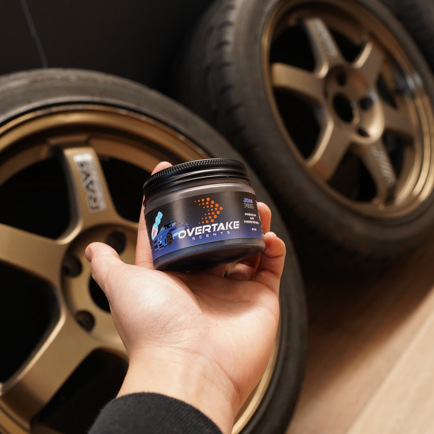 JDM Fizz gel car air freshener held in hand in front of bronze aftermarket alloy wheels — Overtake Scents