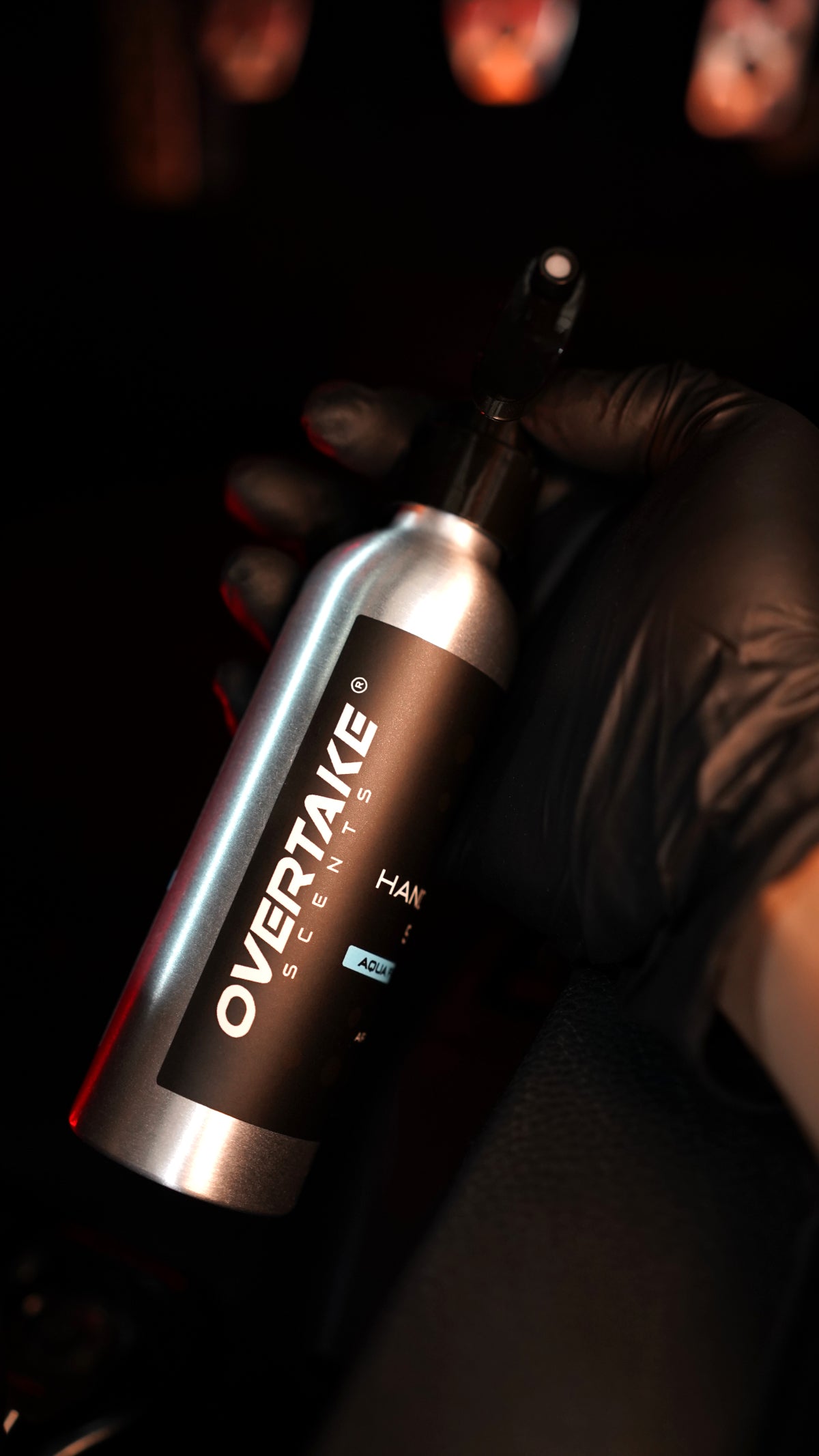 Overtake Handover Spray 150ml held at angle in black-gloved hand, dark background with warm bokeh lighting — Overtake Scents