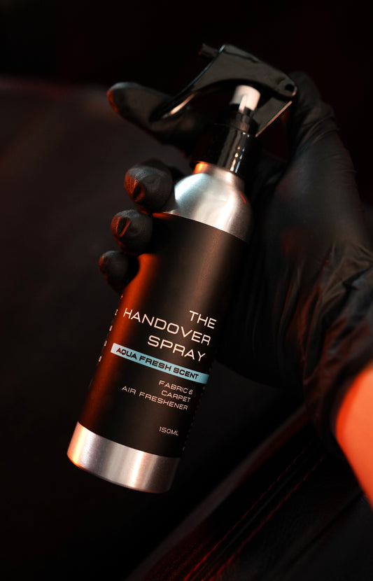 Overtake Handover Spray 150ml held in black-gloved hand, dark moody background with warm amber lighting — Overtake Scents