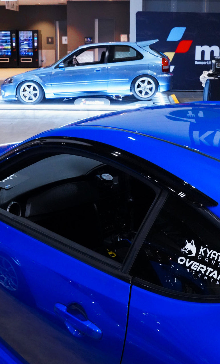 Overtake Scents vent mount displayed in blue Subaru BRZ at indoor car show, collab sticker on rear window — Overtake Scents