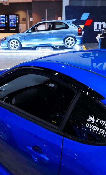 Overtake Scents vent mount displayed in blue Subaru BRZ at indoor car show, collab sticker on rear window — Overtake Scents